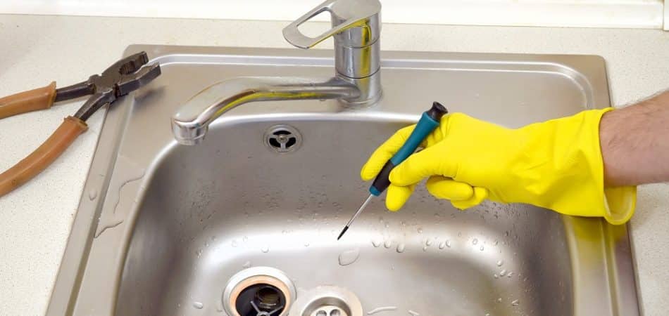 A person wearing a yellow rubber glove uses a screwdriver to work on a kitchen sink drain, tackling clogged drains in Maryville; pliers lie nearby on the countertop.