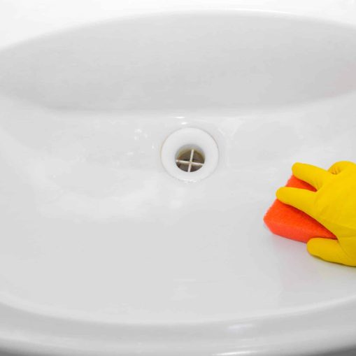A person wearing a yellow rubber glove cleans a white sink with an orange sponge, showcasing the importance of regular drain cleaning in Knoxville.