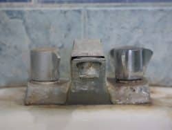 Close-up of an old, corroded bathroom faucet with two metal knobs and visible rust, white buildup on faucets in Maryville, and mineral deposits on the surface and sink area.