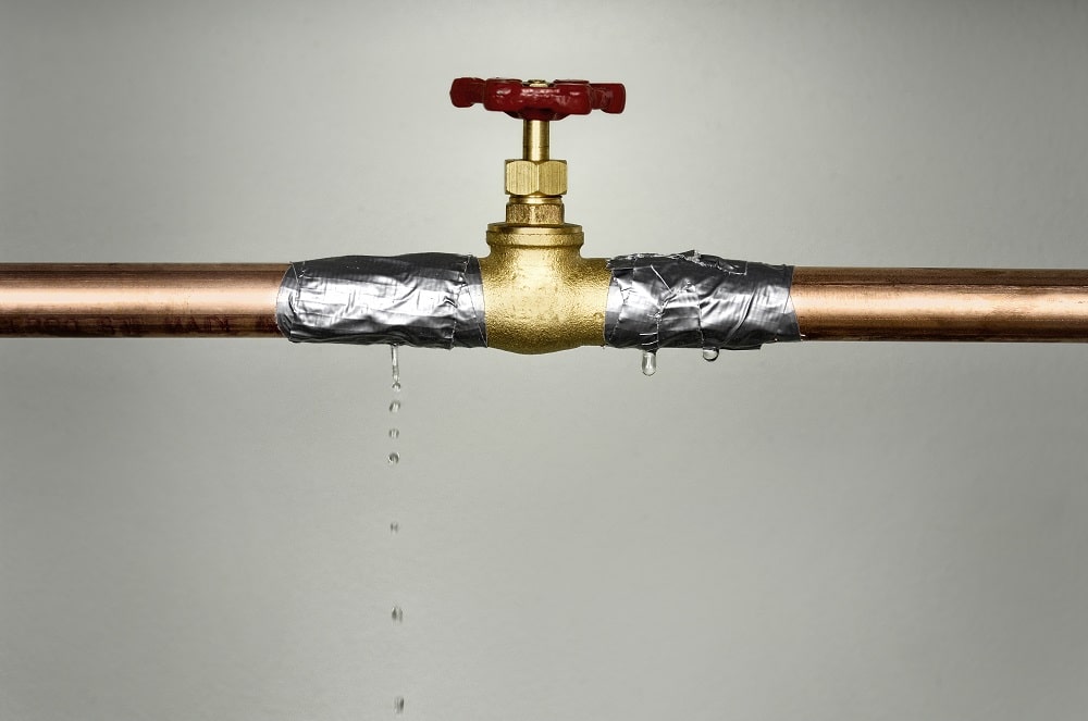 A brass valve on a copper pipe wrapped with duct tape struggles against high water pressure, leaking droplets onto a plain background.