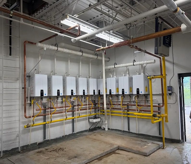 A row of six wall-mounted tankless water heaters connected by copper and yellow gas pipes in a utility room with exposed ceiling.