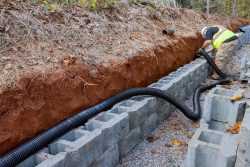 A worker in a safety vest installs a black drainage pipe behind a retaining wall made of concrete blocks in a trench, showcasing expert installation of Drainage Systems in Knoxville.