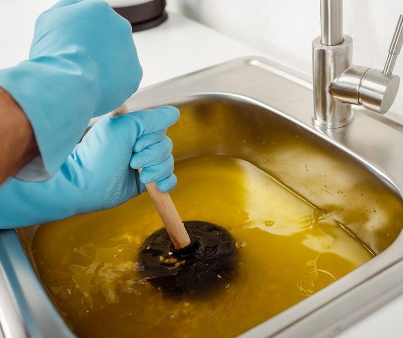 A person wearing blue gloves uses a plunger for drain cleaning in Knoxville, working on a stainless steel sink filled with murky yellow water.