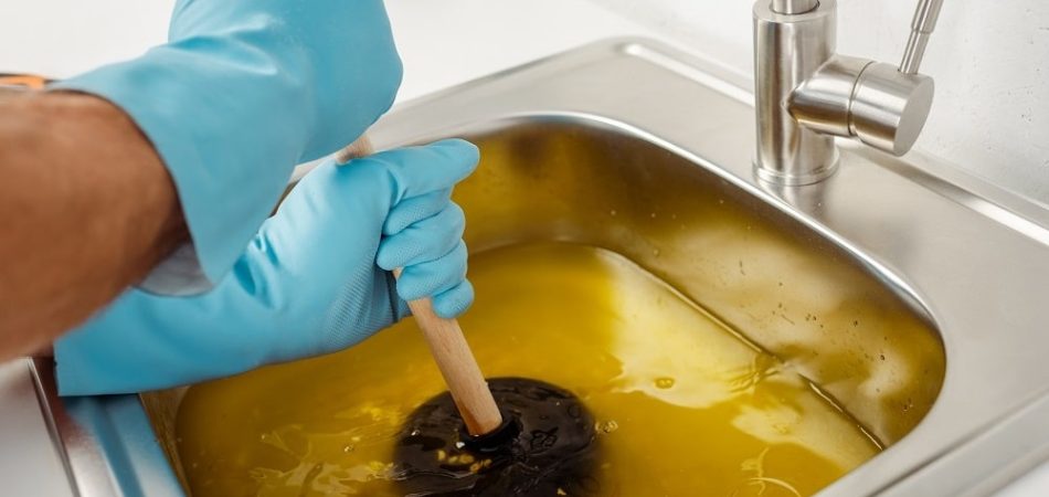 A person wearing blue gloves uses a plunger to unclog kitchen sink drains filled with yellowish water.