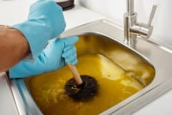 Person wearing blue gloves uses a plunger in a stainless steel sink filled with murky yellow-brown water.