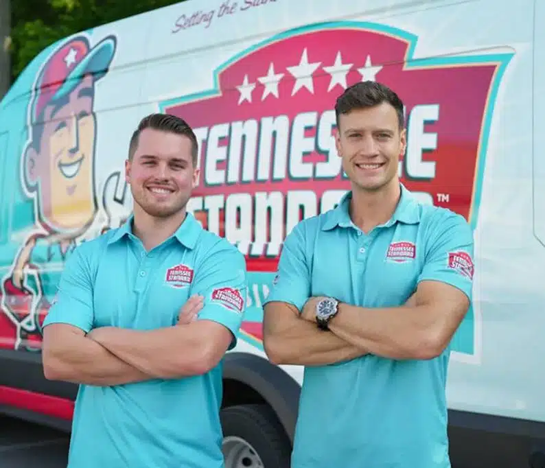 Bobby and Kelton Balka wearing light blue shirts stand smiling with arms crossed in front of a van displaying the Tennessee Standard logo and colorful graphics.
