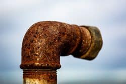 Close-up of a rusty, corroded metal pipe with an elbow joint highlights the impact of corrosion inside plumbing systems, set against a blurred light blue and gray background.