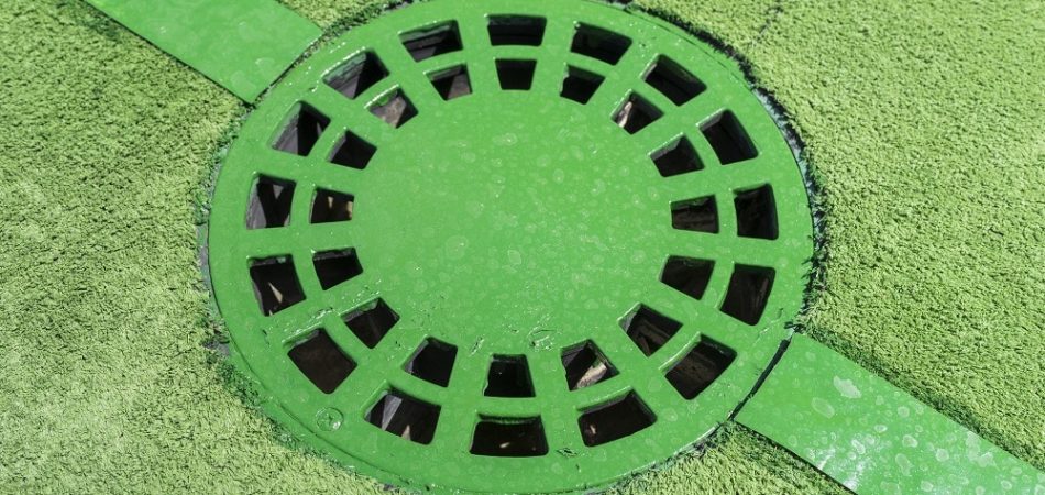A close-up of a green circular drainage grate set into a green textured surface, intersected by two green strips—an ideal spot for outdoor drain unclogging.
