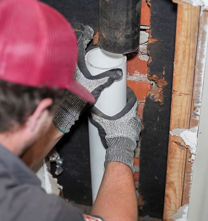 Person wearing gloves and a red hat installs or adjusts a white PVC pipe inside a wall with exposed brick and wood framing.