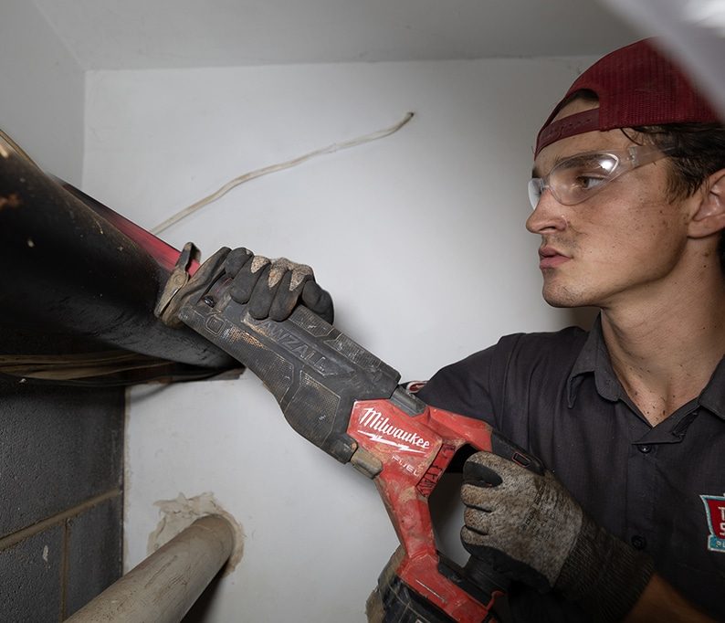 Person wearing safety goggles and gloves uses a reciprocating saw to cut through a black pipe in a small indoor space.