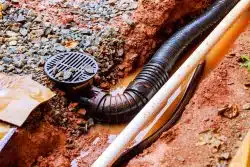 Black plastic drainage pipe and white conduit partially buried in wet soil and gravel, with muddy water pooling around them—showcasing typical Drainage Systems in Knoxville.