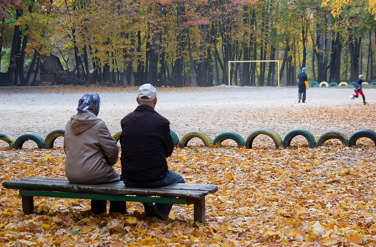 Two people sit on a bench in a park covered with autumn leaves, watching children play near a soccer goal in the background.