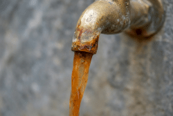 A close-up of a rusty metal faucet with brown, murky water flowing out, suggesting corrosion inside plumbing systems and indicating contaminated or unclean water.