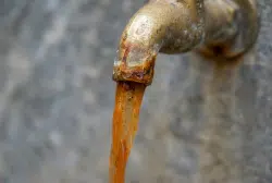 A close-up of a rusty metal faucet with brown, murky water flowing out, suggesting corrosion inside plumbing systems and indicating contaminated or unclean water.