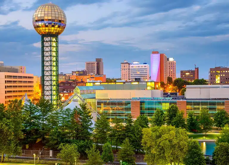 Knoxville, Tennessee skyline at dusk featuring the Sunsphere tower and surrounding downtown buildings with green trees in the foreground.