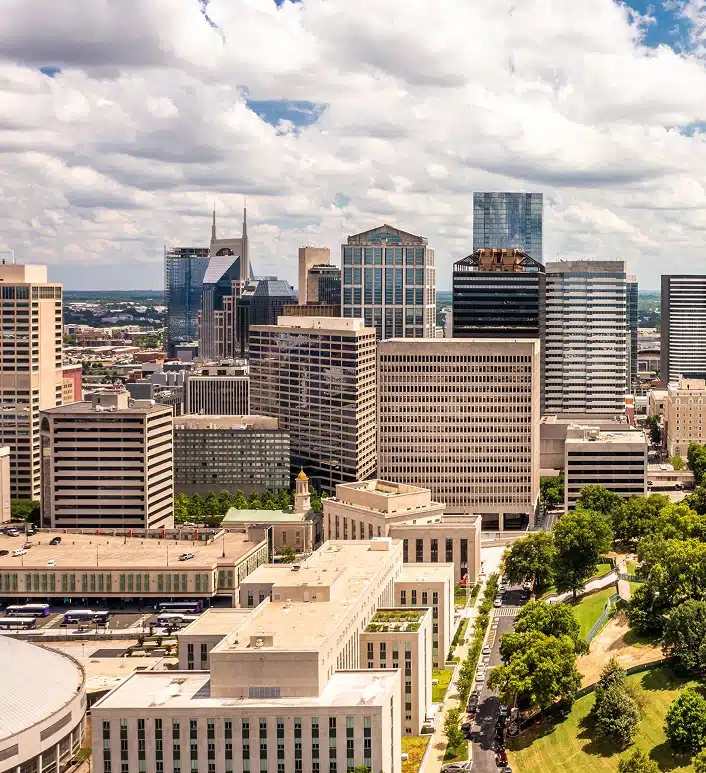 Aerial view of a downtown cityscape with high-rise office buildings, a state capitol building, and tree-lined streets under a partly cloudy sky.