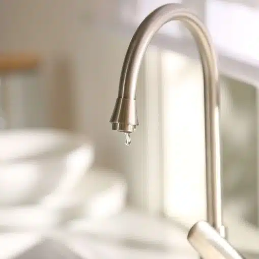 A close-up of a kitchen faucet with a single water droplet about to fall, with blurred dishes and sunlight in the background.