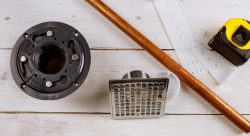 Plumbing parts for shower drain installation, including a copper pipe, a drain grate, a measuring tape, and white plastic brackets, are neatly arranged on a wooden surface.