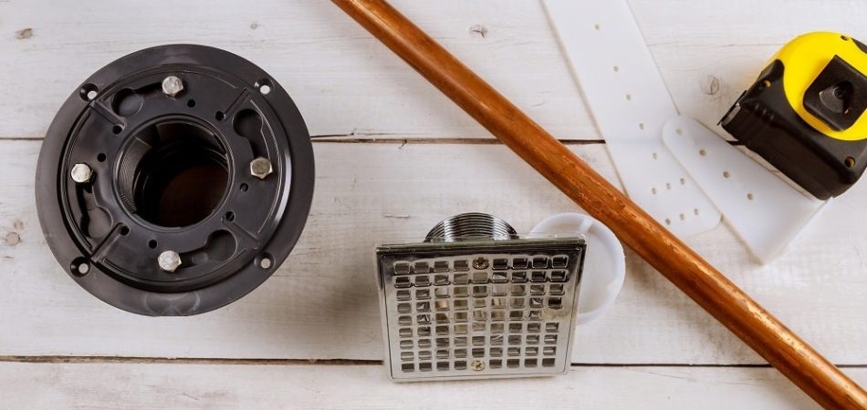 Plumbing parts for shower drain installation, including a copper pipe, a drain grate, a measuring tape, and white plastic brackets, are neatly arranged on a wooden surface.