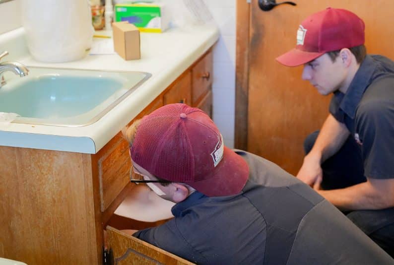 Two workers in uniforms and red caps inspect the area under a bathroom sink, with one kneeling and the other observing.