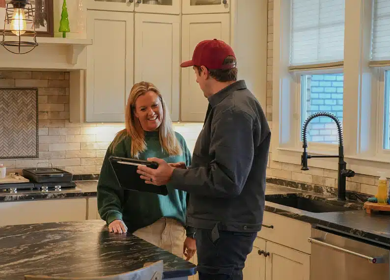 A woman and a man stand in a kitchen by a marble island; the man holds a tablet and they appear to be having a discussion.