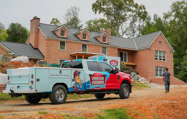 A Tennessee Standard Plumbing truck is parked in front of a large brick house under construction; a worker is walking toward the building.