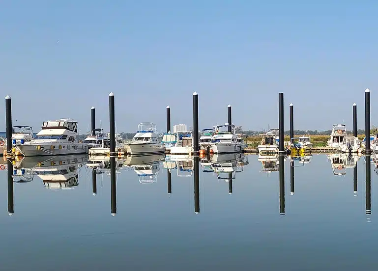 Row of boats docked at a calm marina with their reflections mirrored in the still water under a clear sky.