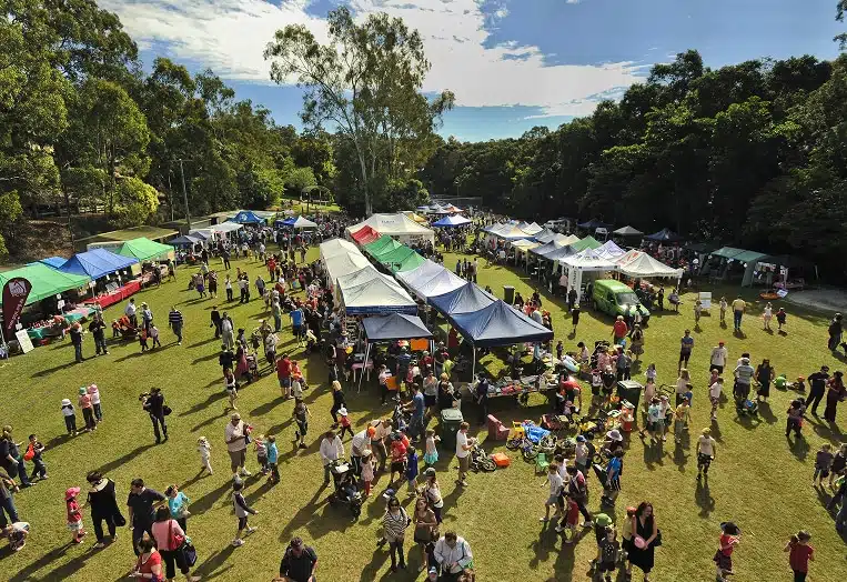 Aerial view of an outdoor community festival with colorful tents, vendor booths, and crowds of people gathered on a green field surrounded by trees.