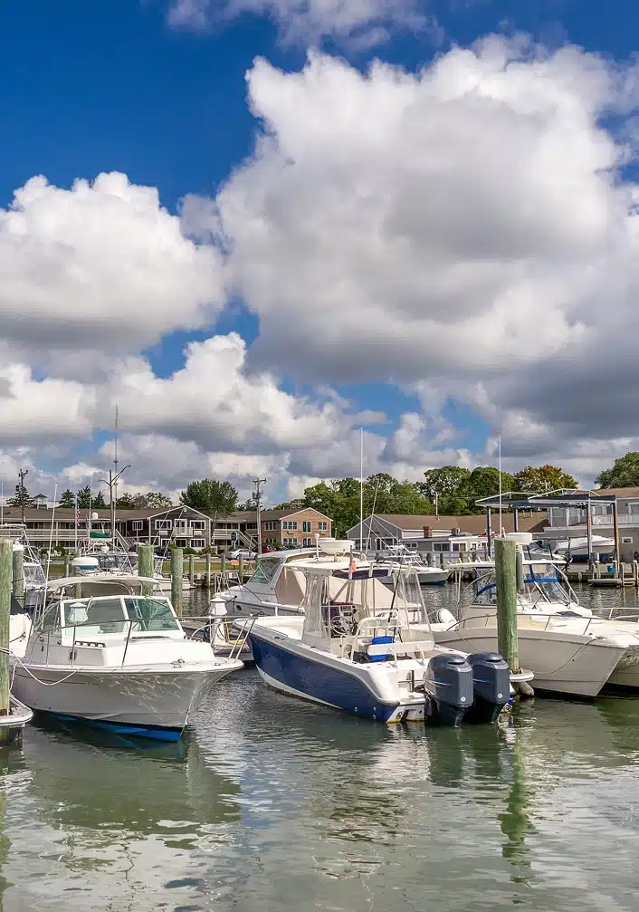 Several motorboats moored at a marina dock with waterfront buildings in the background under a partly cloudy sky.