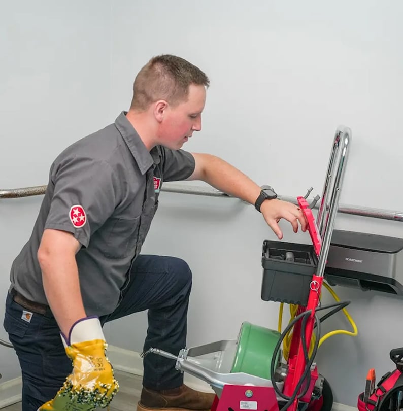 A technician in a gray uniform kneels next to cleaning equipment and adjusts controls on a red floor cleaning machine in a room with white walls.