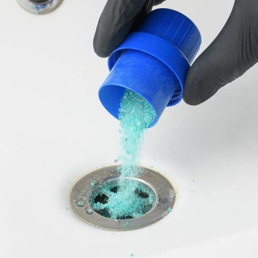 A hand wearing a black glove pours blue drain cleaner powder into a sink drain.