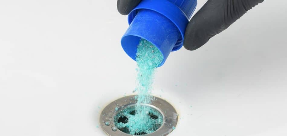 A hand wearing a black glove pours blue drain cleaner powder into a sink drain.