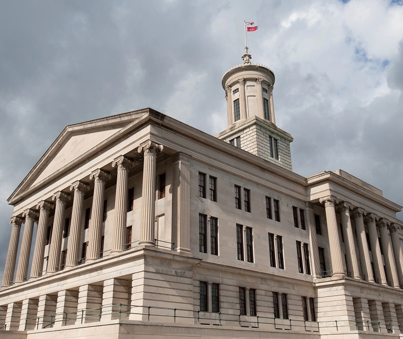 A neoclassical government building with tall columns and a central tower, topped by a flag against a cloudy sky.