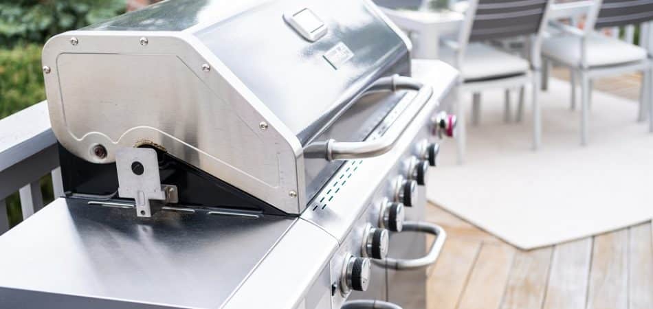 A stainless steel gas grill, connected to a gas line for an outdoor kitchen in Maryville, sits on a wooden deck with a patio table and chairs in the background. Red, white, and blue bunting hangs on the railing.