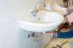 A person repairs or inspects the bathroom sink drain under a wall-mounted white bathroom sink with a chrome faucet.