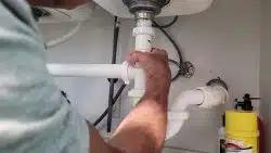 Person installing kitchen sink drain by tightening a white PVC pipe connector under the sink with both hands; cleaning supplies are visible nearby.