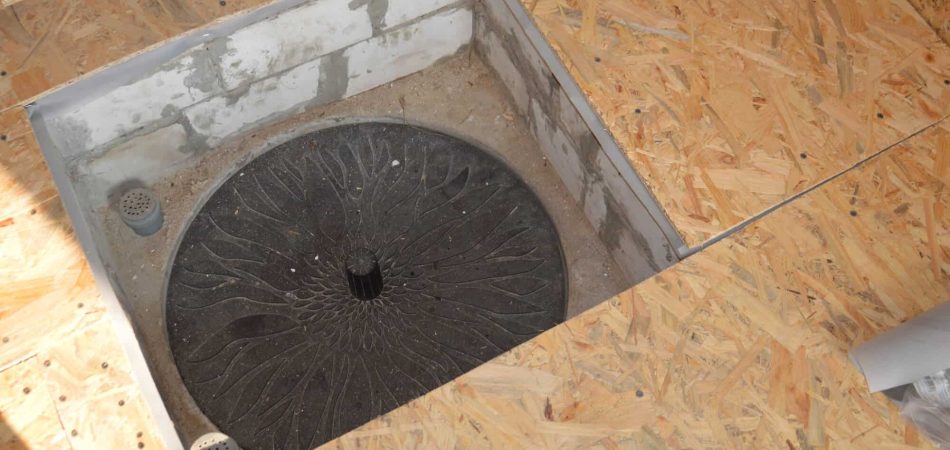 A circular black drain cover, designed to hide sump pump access, is installed in a square cutout within an unfinished oriented strand board (OSB) floor.