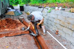 A man installs a black drainage pipe in a trench next to a retaining wall, showcasing effective Drainage Systems in Knoxville, using tools and surrounded by construction materials.