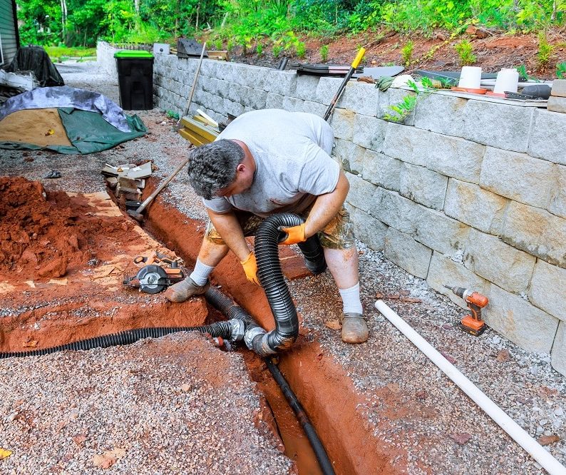 A man installs a black drainage pipe in a trench next to a retaining wall, showcasing effective Drainage Systems in Knoxville, using tools and surrounded by construction materials.