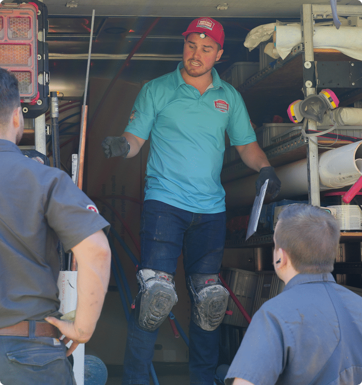 Kelton Balka, CEO of Tennessee Standard Plumbing, wearing a blue shirt and red cap, steps down from a truck with a clipboard in hand, facing two people nearby.