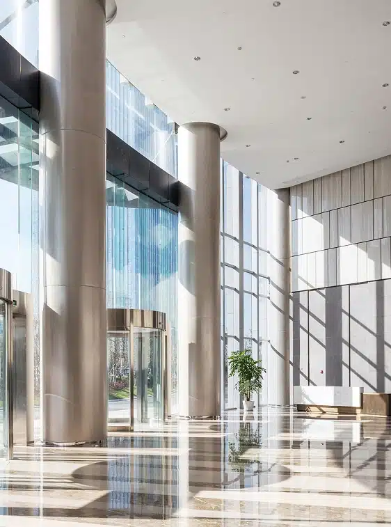 Modern office lobby with tall glass windows, large columns, polished floors, a revolving door, a potted plant, and a white bench. Sunlight casts shadows on the floor and walls.