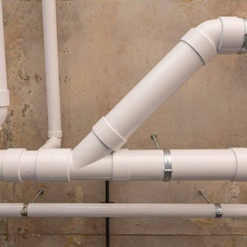 White PVC plumbing pipes arranged against a concrete wall, connected with elbows and metal brackets for support, may indicate improperly slope drain pipes affecting efficient water flow.