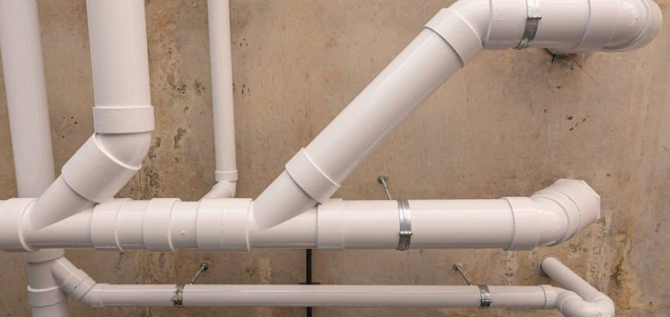 White PVC plumbing pipes arranged against a concrete wall, connected with elbows and metal brackets for support, may indicate improperly slope drain pipes affecting efficient water flow.