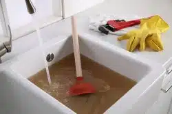 A kitchen sink filled with murky brown water, a plunger inside, water running from the tap, and rubber gloves with a pipe wrench on the counter—perfect candidates for Professional Drain Cleaning in Maryville.
