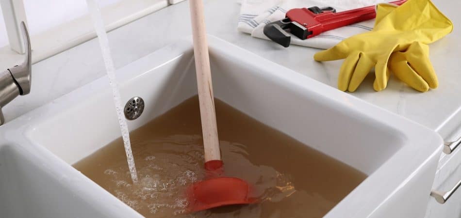 A kitchen sink filled with dirty brown water signals repeated drain clogs, with a plunger submerged in the water, gloves and a pipe wrench on the counter, and water still running from the faucet.