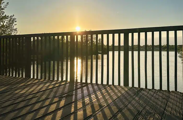 Sunset view from a wooden deck, with shadows cast by railings. The sun reflects on a calm lake, surrounded by silhouetted trees, creating a serene mood