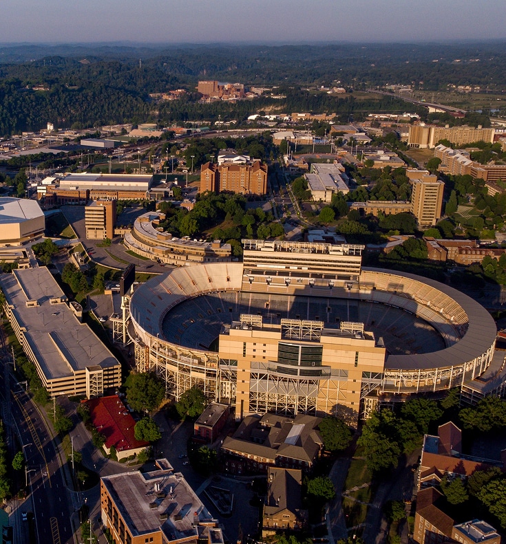 Aerial view of a large, empty football stadium surrounded by buildings and trees, with hills and cityscape in the background.