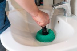 Person performing drain cleaning in Knoxville, using a green plunger in a white bathroom sink with water running from the faucet.