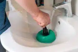 Person performing drain cleaning in Knoxville, using a green plunger in a white bathroom sink with water running from the faucet.