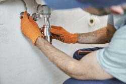 A person wearing orange gloves repairs a pipe under a sink—perfect for anyone searching for a Plumber Near Me in Oak Ridge.